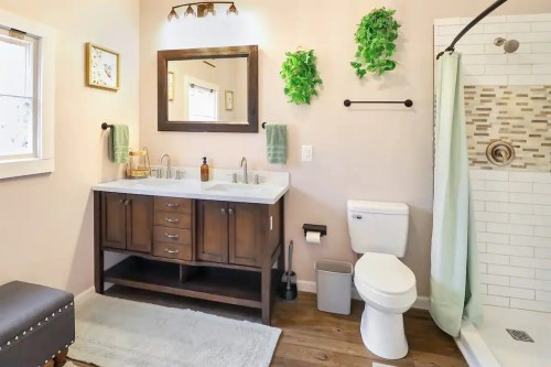 A modern bathroom featuring a double sink vanity, shower, toilet, and greenery accents on the walls.