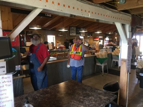 Two men inside a rustic store, one in overalls and the other in a security vest, with snacks and decor in the background.