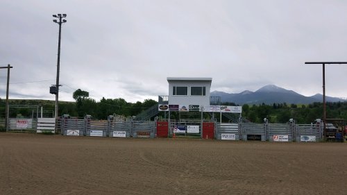 A rodeo arena with a grandstand, surrounded by mountains under a cloudy sky. Fenced area with sponsor banners visible.