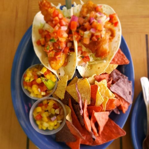 Two fish tacos topped with pico de gallo on a blue plate, accompanied by colorful tortilla chips and salsa.
