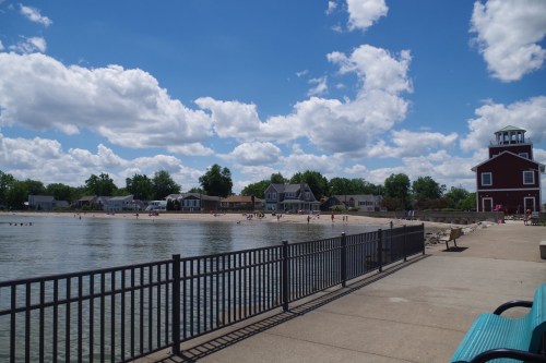A sunny lakeside scene with a sandy beach, houses, and fluffy clouds in the sky, viewed from a paved walkway.