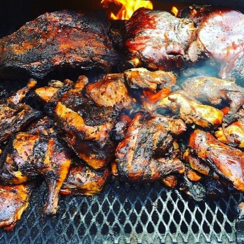 A close-up of various pieces of smoked meat, including chicken and pork, sizzling on a grill with flames in the background.