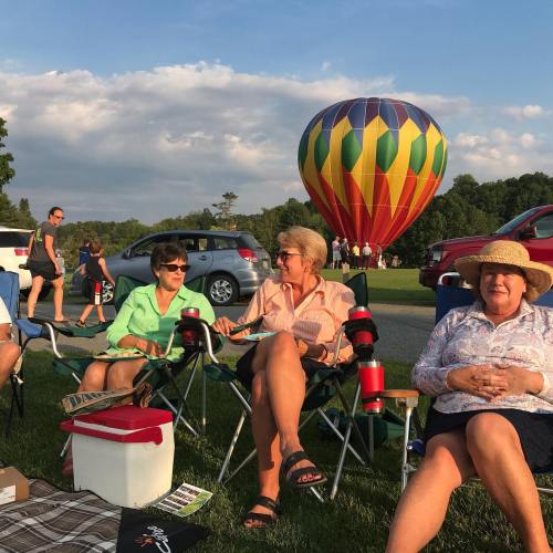 A group of people sitting on lawn chairs at a park, with a colorful hot air balloon in the background.