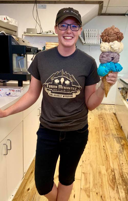 A smiling person in a cap holds a colorful ice cream cone with four scoops, standing in an ice cream shop.