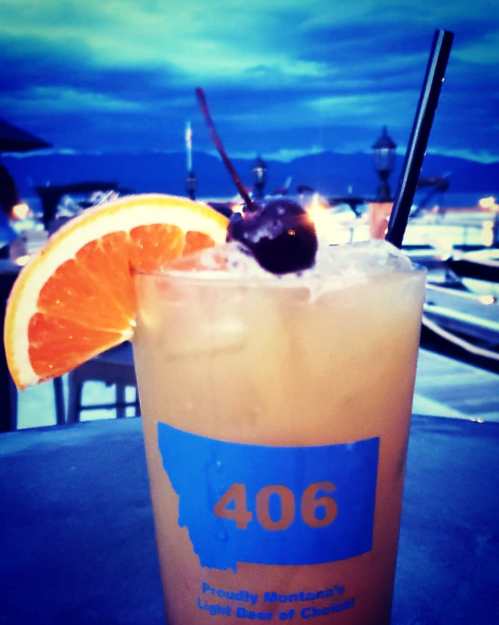 A cocktail in a glass with an orange slice and cherry, set against a moody sky and outdoor seating.