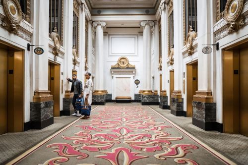 Two people stand in an ornate hotel lobby with gold accents and a patterned carpet, waiting for an elevator.