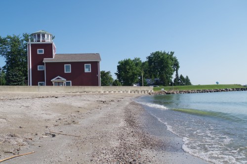 A red building near a sandy beach, with calm water and green trees under a clear blue sky.