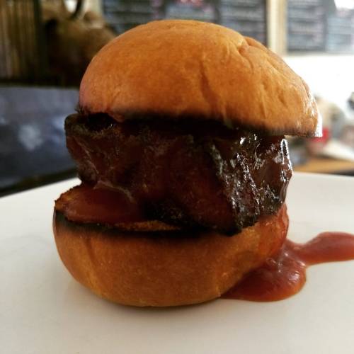 A close-up of a small, toasted bun filled with a piece of grilled meat and drizzled with barbecue sauce.