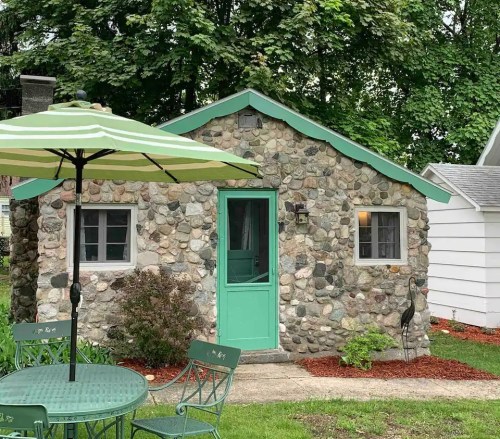A small stone cottage with a green roof and door, surrounded by greenery and a patio set with an umbrella.