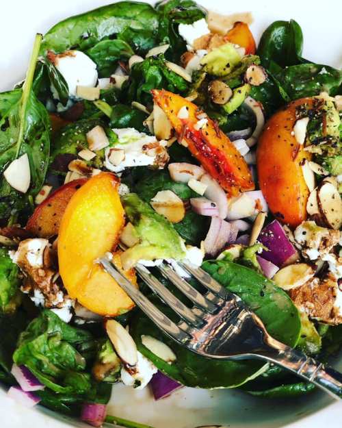A vibrant salad with spinach, peaches, goat cheese, almonds, and red onion, served in a white bowl with a fork.