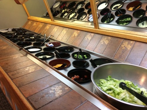 A salad bar with various toppings in black bowls and a large bowl of lettuce in the foreground.