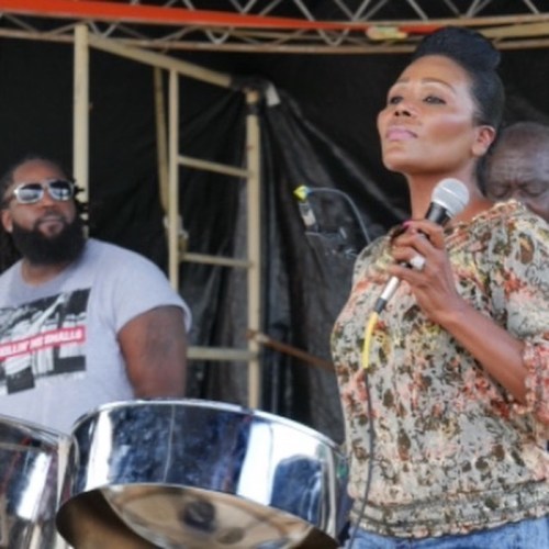 A woman sings passionately into a microphone while a man plays steel drums in the background at an outdoor event.
