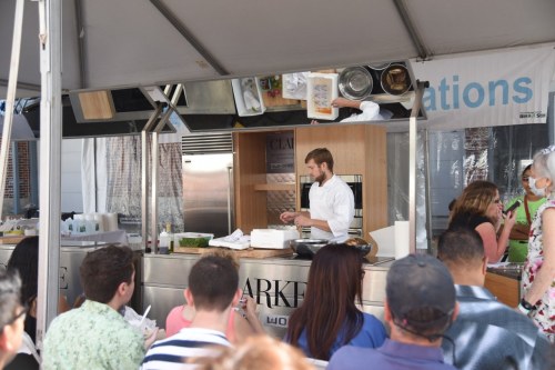 A chef demonstrates cooking techniques at a food event, with an audience watching attentively from the front.