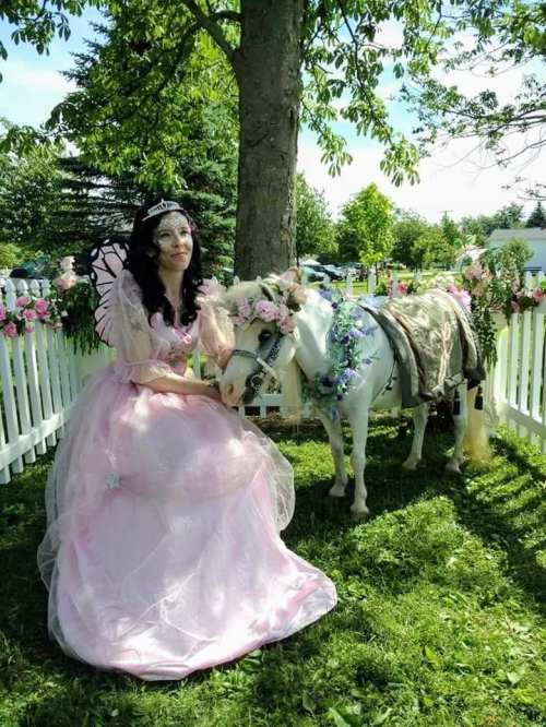 A woman in a pink fairy costume sits beside a small decorated pony in a garden with flowers and greenery.