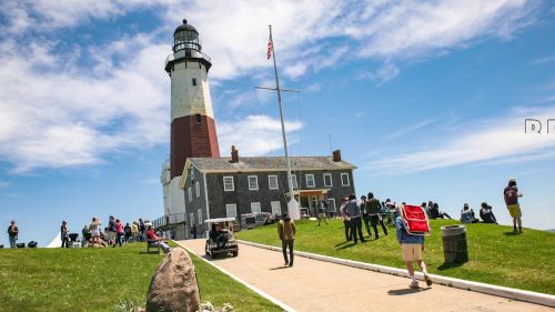 A lighthouse stands tall against a blue sky, with visitors walking on a path and enjoying the scenic view.