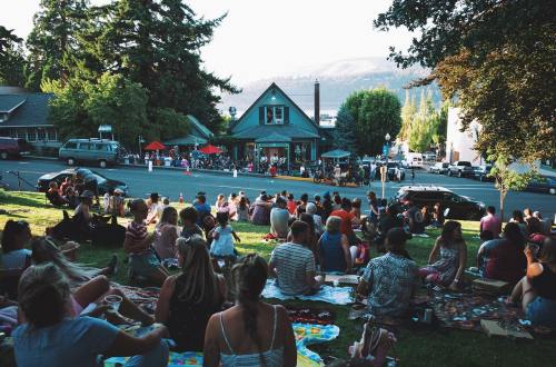 A crowd gathers on a grassy area, enjoying a sunny day near a lake, with trees and a building in the background.