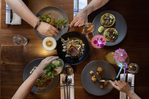 A top-down view of a dining table with various colorful dishes and drinks, hands reaching for food.