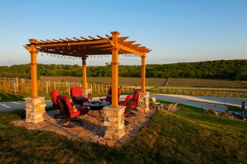 A cozy outdoor seating area with red chairs under a wooden pergola, overlooking a vineyard and green hills.