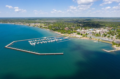 Aerial view of a marina with boats, surrounded by green trees and a small town along the shoreline under a blue sky.
