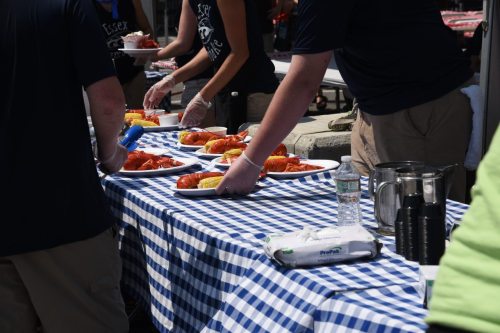 People serving lobster and corn on a blue and white checkered tablecloth at an outdoor event.
