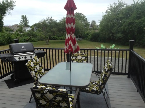 A patio with a glass table, floral chairs, a grill, and a red and white umbrella, overlooking a grassy yard.