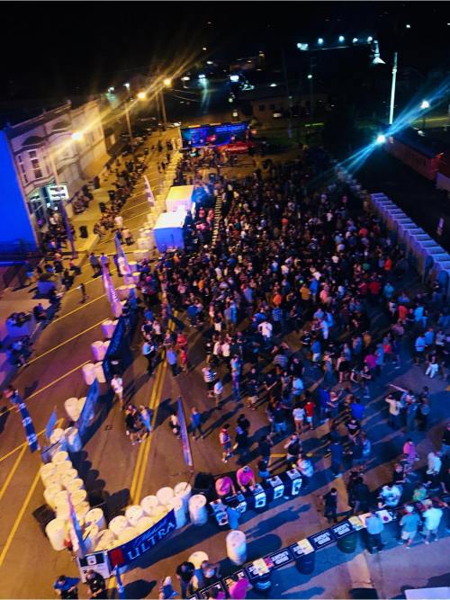 Aerial view of a large crowd gathered at night for an outdoor event, with colorful lights and a stage in the background.