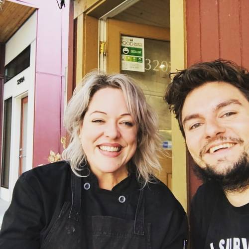 Two smiling individuals pose together outside a restaurant, one in a chef's coat, with a welcoming atmosphere.
