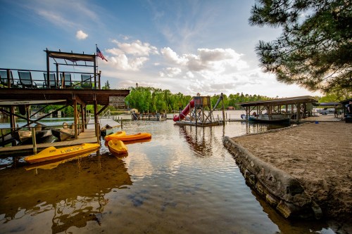 A serene lakeside scene with kayaks, a water slide, and a wooden dock under a partly cloudy sky.