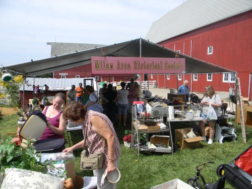 A bustling outdoor market scene under a tent, with people browsing items for sale near a red barn.