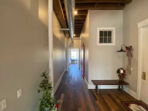 A long hallway with wooden floors, a bench, and a small decorative tree, leading to a bright room at the end.