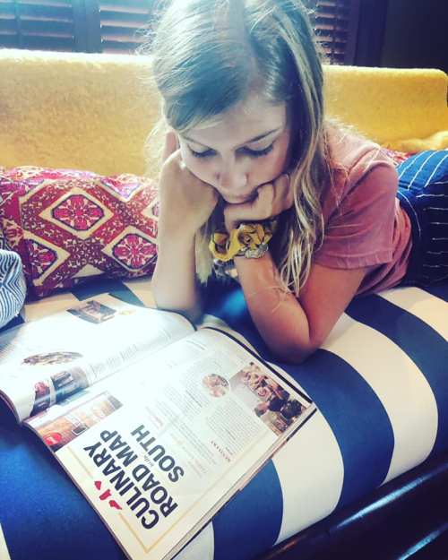 A young woman reading a magazine while lounging on a striped couch, resting her chin on her hands.