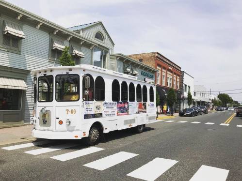 A white trolley bus parked on a street lined with shops and trees, with a clear sky overhead.