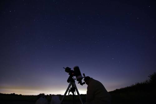 A person observes the night sky through a telescope, surrounded by stars in a dark landscape.