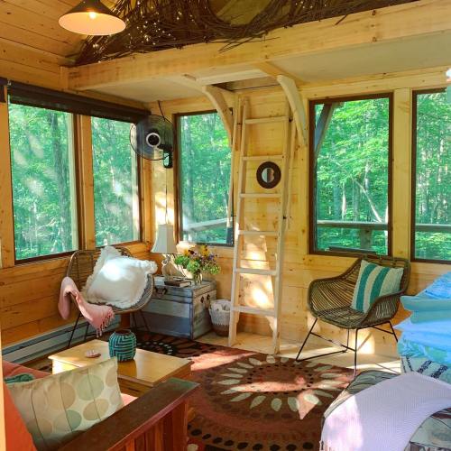 Cozy interior of a cabin with large windows, wooden decor, a ladder, and comfortable seating surrounded by greenery.
