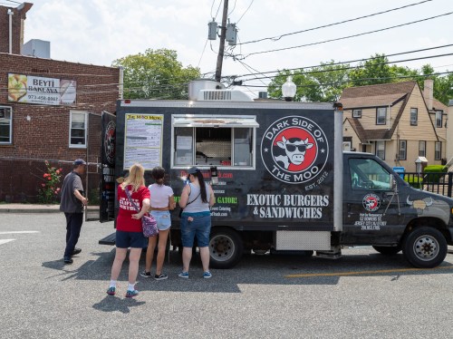 A food truck named "Dark Side of the Moo" serves exotic burgers and sandwiches, with customers waiting in line.
