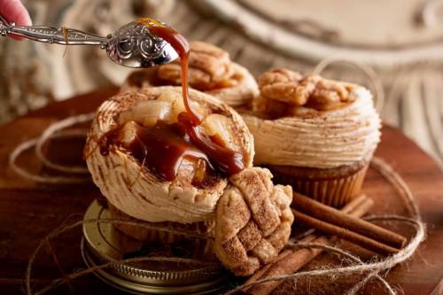 A spoon drizzles caramel over beautifully decorated cupcakes with cinnamon and apple filling on a rustic wooden surface.