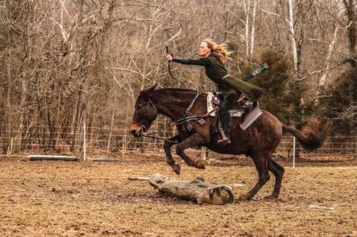 A woman in a green cloak rides a horse, drawing a bow while leaping over a log in a wooded area.
