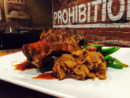 A plate featuring glazed ribs, green beans, and a serving of mashed sweet potatoes, with a brick wall in the background.