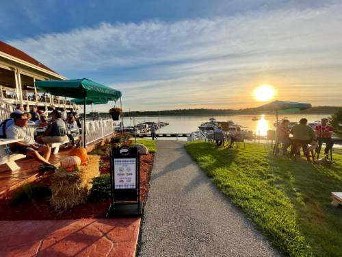 A scenic lakeside view at sunset, with people dining outdoors and boats docked nearby.