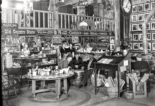 A vintage general store interior with shelves of goods, a round table, and customers chatting. Black and white photo.