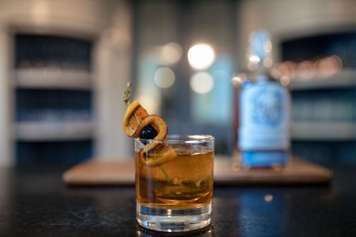 A cocktail with orange and cherry garnishes in a glass, with a bottle of liquor blurred in the background.