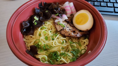A bowl of noodles topped with grilled meat, sliced egg, pickled vegetables, and black mushrooms, with green onions sprinkled on top.