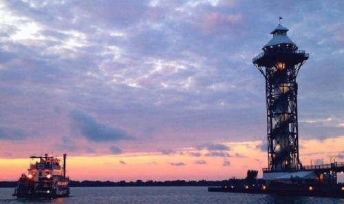 A boat sails near a tall observation tower against a colorful sunset sky with scattered clouds.