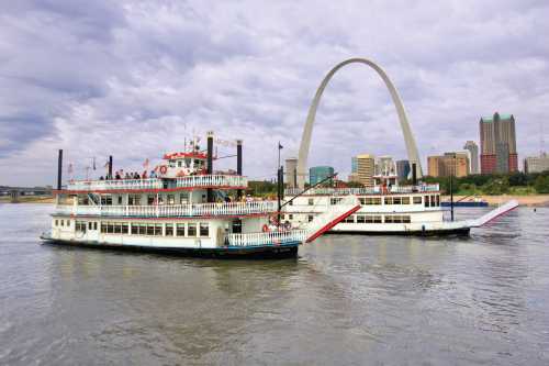 Two riverboats navigate the water near the Gateway Arch and city skyline under a cloudy sky.