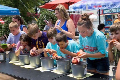 Children compete in an apple-eating contest, focused on their buckets, with adults supervising in a festive outdoor setting.
