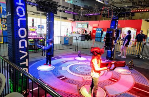 Two children play in a colorful arcade area, using virtual reality guns on a circular platform with lights.