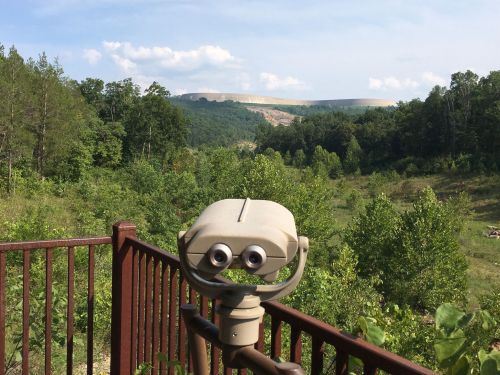 A viewing telescope overlooks a lush green valley with distant hills under a clear blue sky.