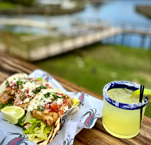 Three fish tacos topped with pico de gallo and cheese, served with a lime, alongside a margarita on a wooden table.