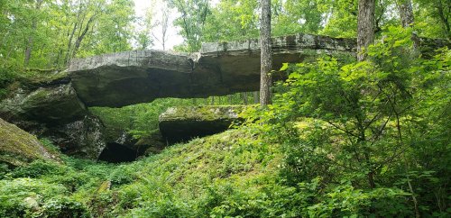A natural stone arch surrounded by lush green trees and foliage in a forested area.