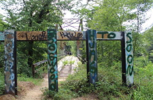 A weathered bridge with graffiti on its supports, surrounded by lush greenery and a dirt path leading across.